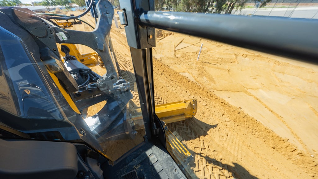 View from a motor grader cab showing the blade shaping soil during road construction work
