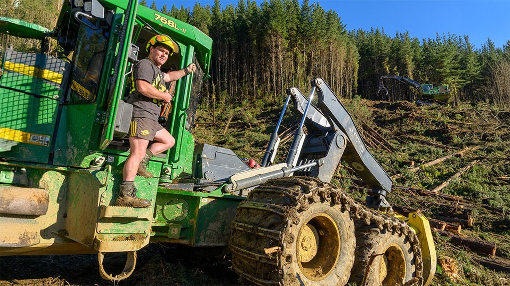 A worker in a green skidder operates the machine in a cleared forest filled with fallen trees.