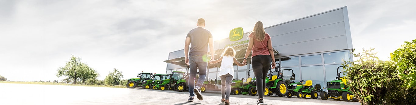 Family walks toward John Deere dealership with green and yellow tractors on display outside.