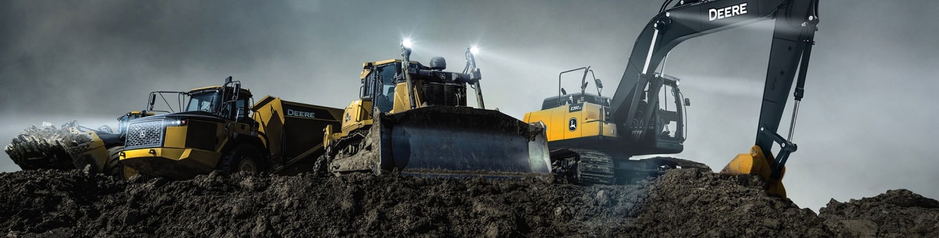 A line up of John Deere construction equipment including an Articulated Dump Truck, Dozer and Excavator, pictured against a dark and stomy sky on a mound of earth 
