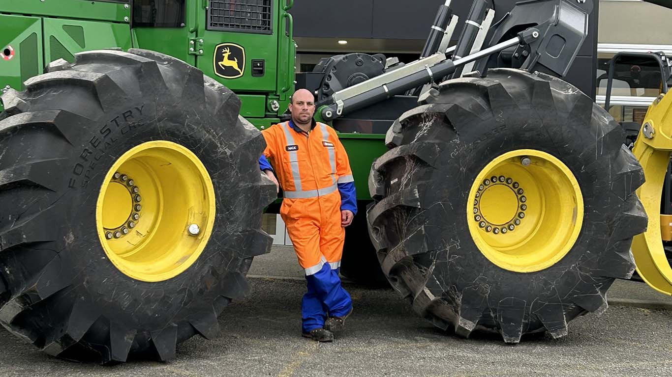 Nick Fargher leaning on the tire of John Deere machine in an orange technician outfit. 