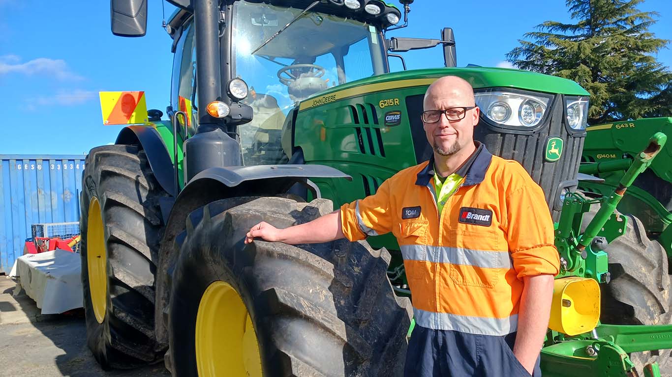 Tom Fyfe wearing a technician's uniform smiling with his hand on the tire of a John Deere tractor outside.