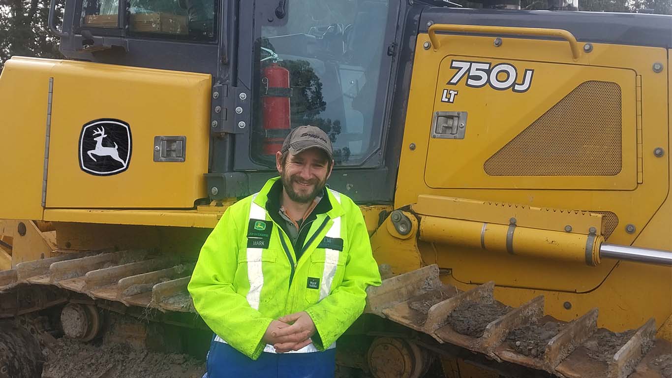 Mark Haughton leaning on the belt tire of a John Deere construction machine wearing a highlighter yellow jacket.