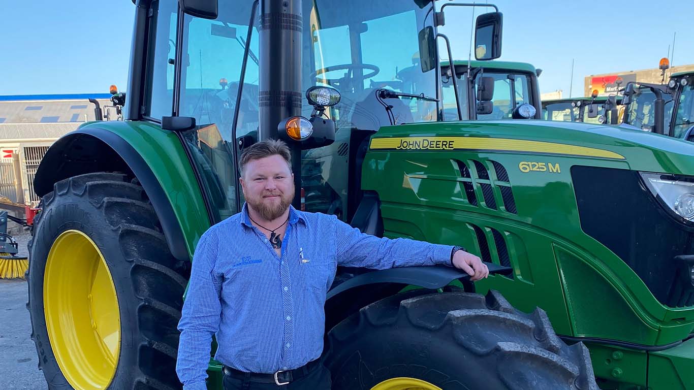 Jimmy O'Donnell leaning on a John Deere tractor with a clear blue sky in the background.