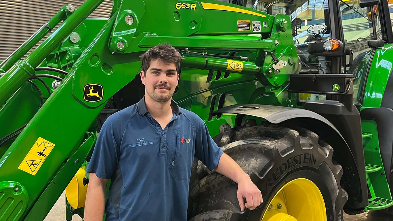 Dylan Bruce leaning on the tire of a John Deere utility tractor inside a manufacturing facility. 