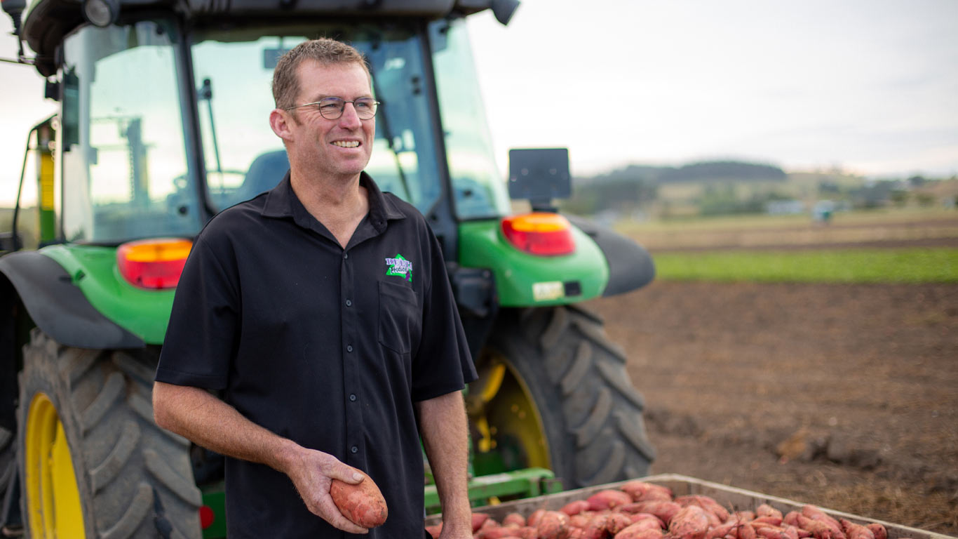 Farmer Evan Paxton standing next to a large wooden crate filled with some of his kumara (sweet potato) crop. He's holding a kumara in his hand. In the background is his John&nbsp;Deere tractor.