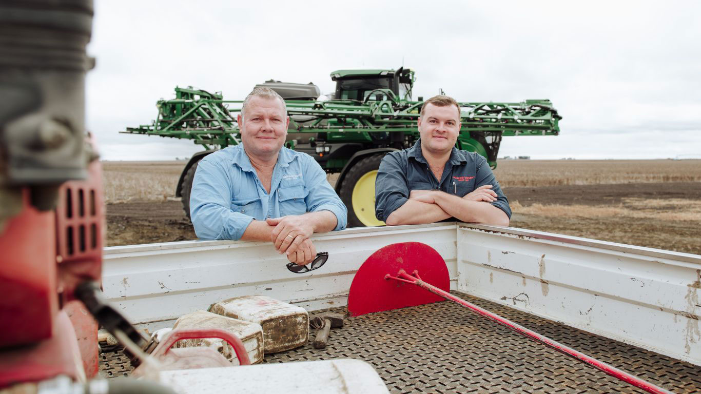 Two men in blue shirts lean on the back of a utility vehicle with farm equipment in the background.