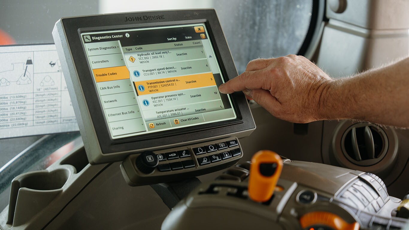 A close-up of a hand interacting with the diagnostics touchscreen inside a John Deere vehicle, showing various troubleshooting codes and system details.