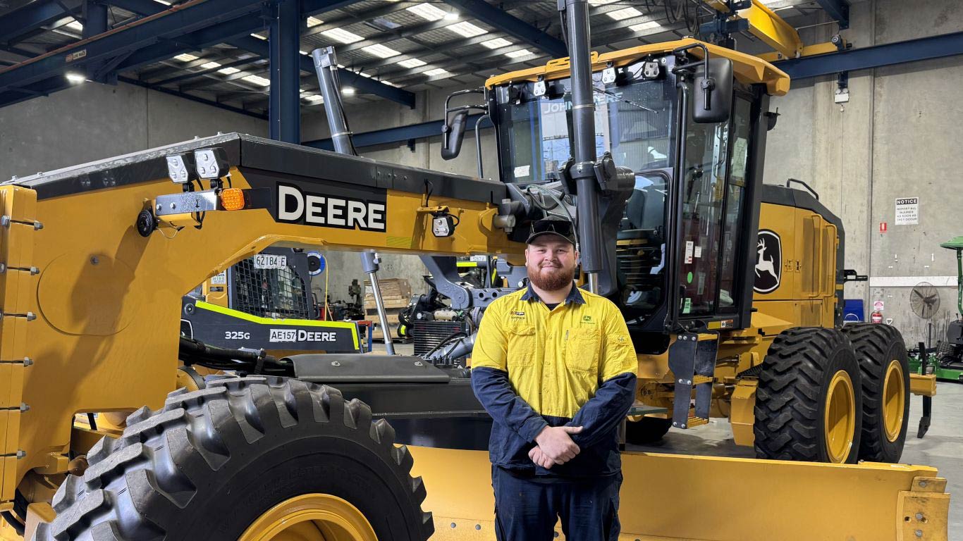 Person standing in front of a John Deere motor grader inside an equipment workshop.