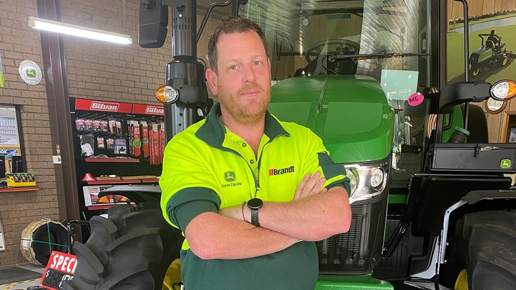 Adam smiling with arms crossed while leaning Deere lawn tractor