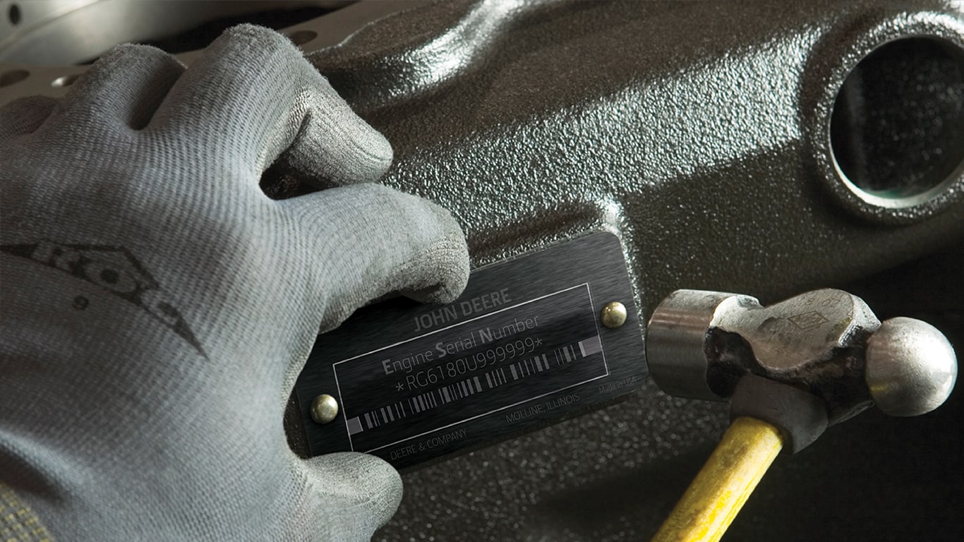 A person wearing a blue glove holding a hammer while installing a John Deere engine serial number plate on an engine