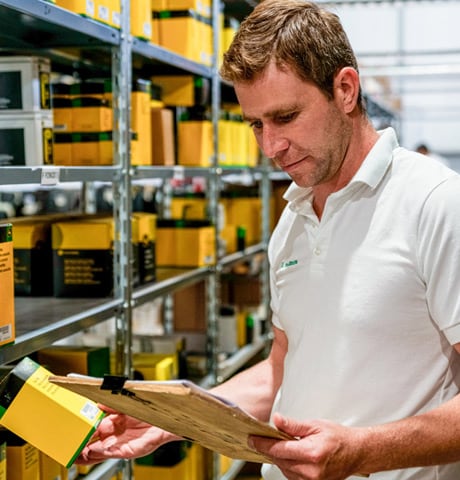 Man in a white polo shirt holding a clipboard examines parts in a warehouse