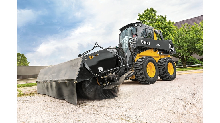 A 320G Skid Steer with broom attachment sweeps grass and dirt off a road.