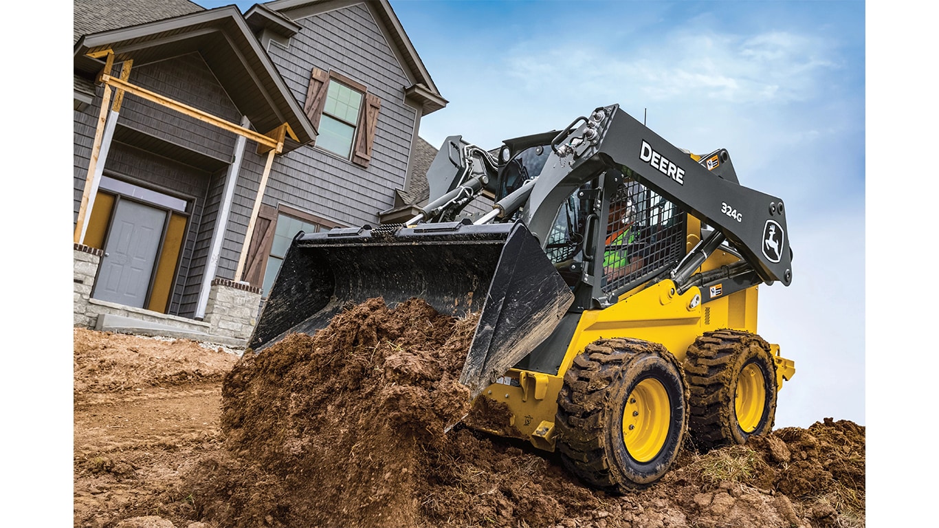 A 324G Skid Steer dumps dirt at a worksite in front of a house.
