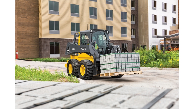 A 324G Skid Steer with pallet attachment transporting bricks in front of a building worksite.