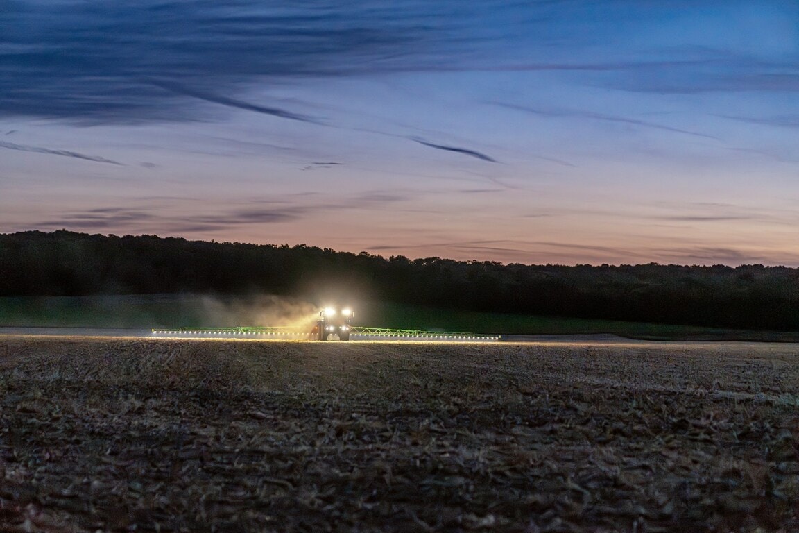John Deere 500R self‑propelled sprayer operating at dusk with LED lights on, wide boom applying crop protection across a field.