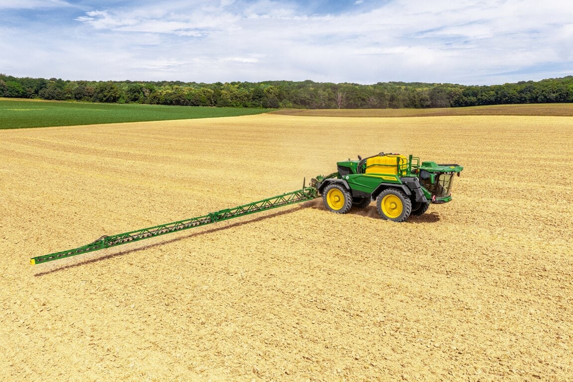John Deere 500R self‑propelled sprayer operating in a wide harvested field, long spray boom extended for crop protection.