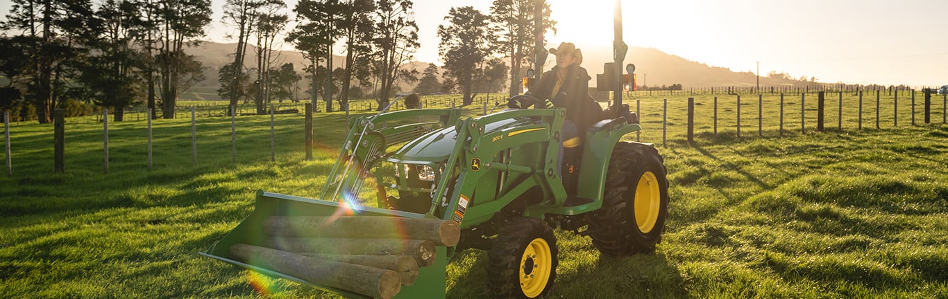 Person drives green tractor with logs in sunny field, trees and hills in background.