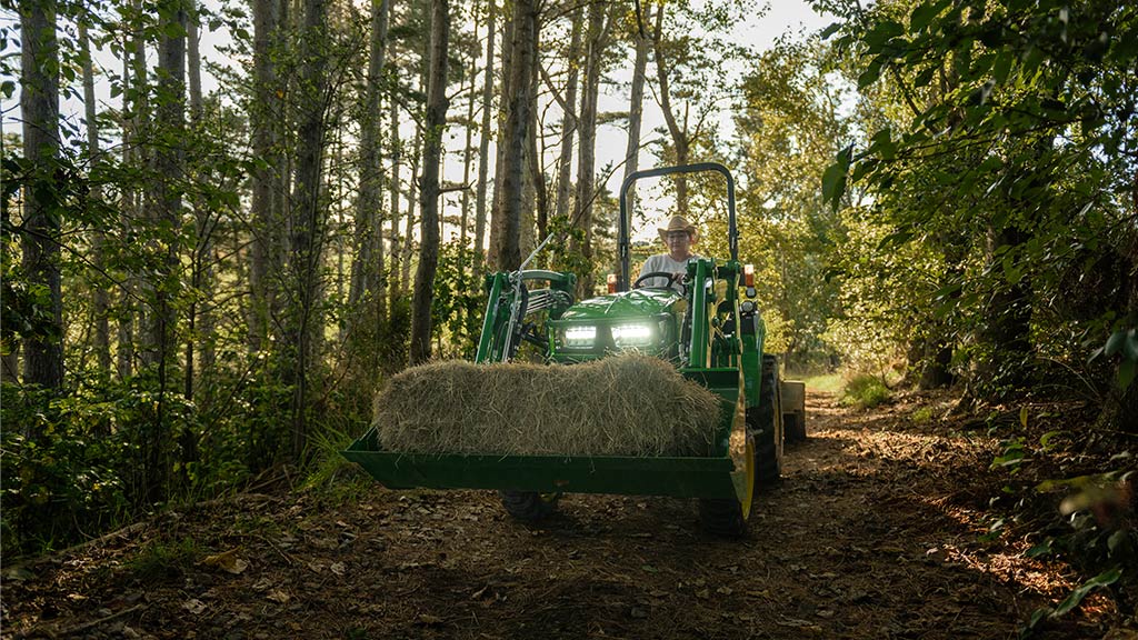 Tractor hauling hay bale on forest path with sunlight filtering through trees.