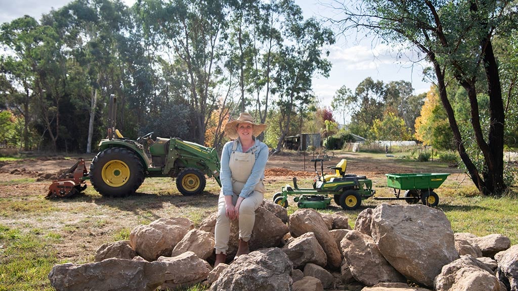 Person in overalls and hat sitting on rocks at farm with tractors and trees in background.