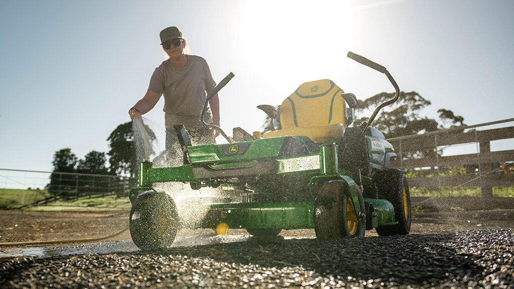 Person sprays green and yellow lawnmower with hose on sunny day.