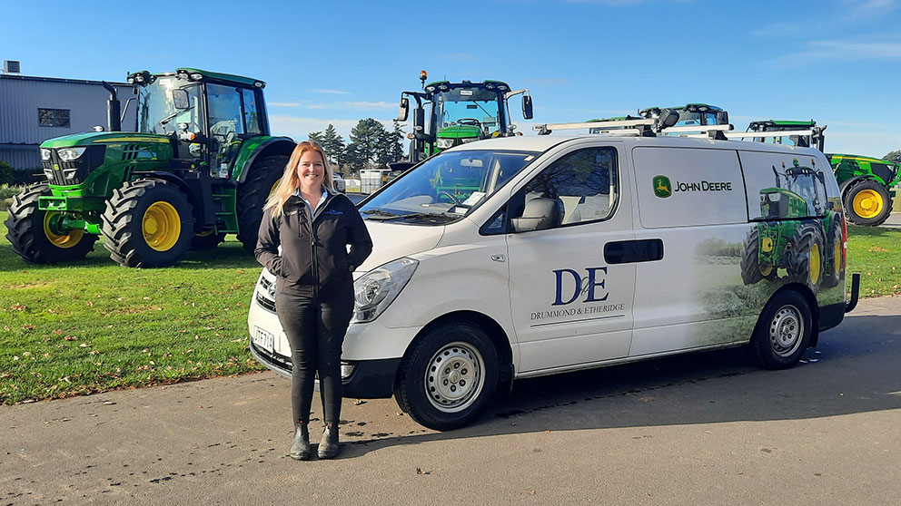 Parts Apprentice Deena Martin, stands in front of a Drummond & Etheridge van with John Deere tractors behind it.