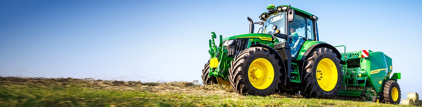 Green John Deere tractor towing a baler across a harvested field under a clear blue sky.