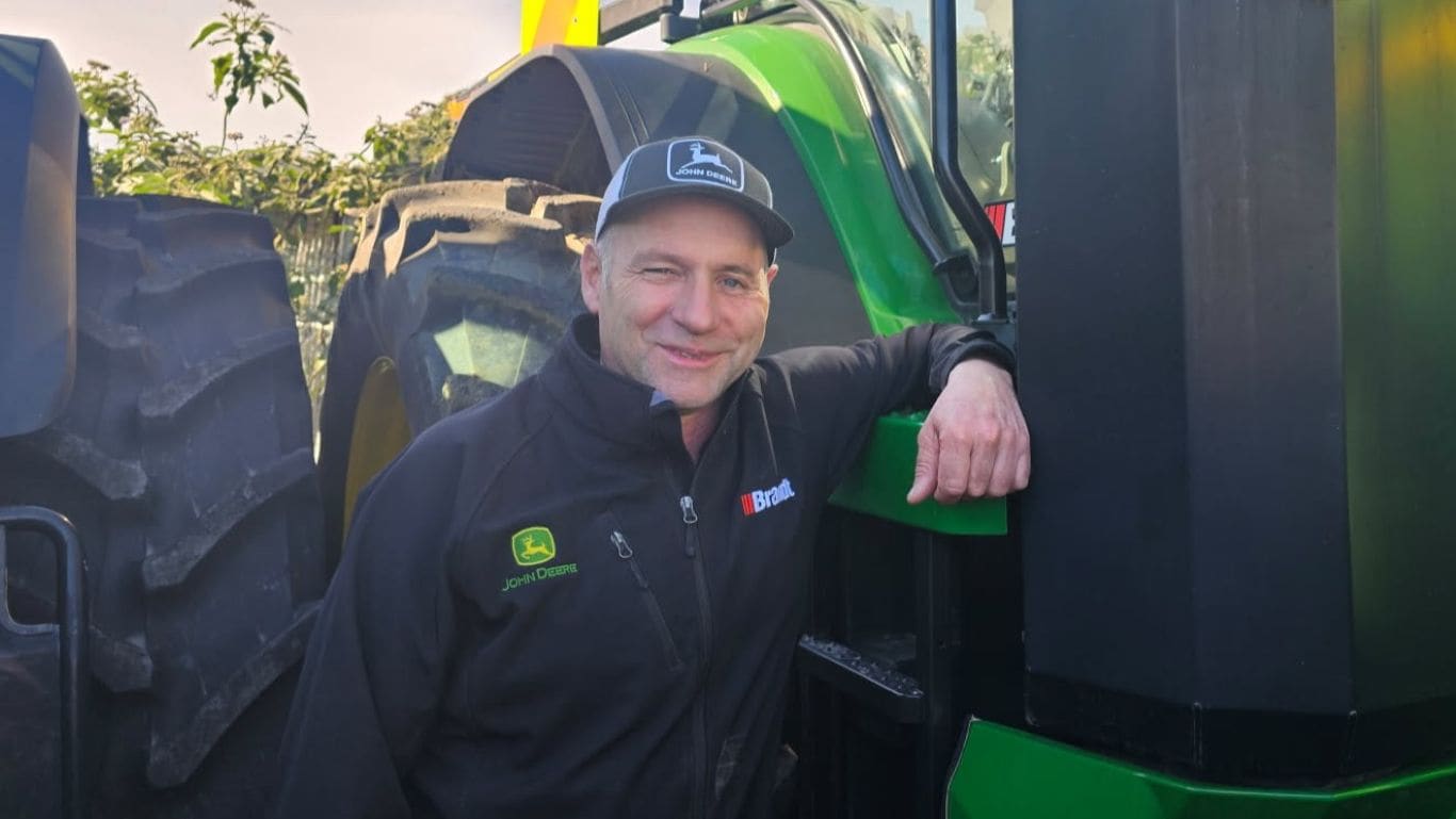 A man wearing a black jumper and a baseball cap leaning on a large green agricultural machine.