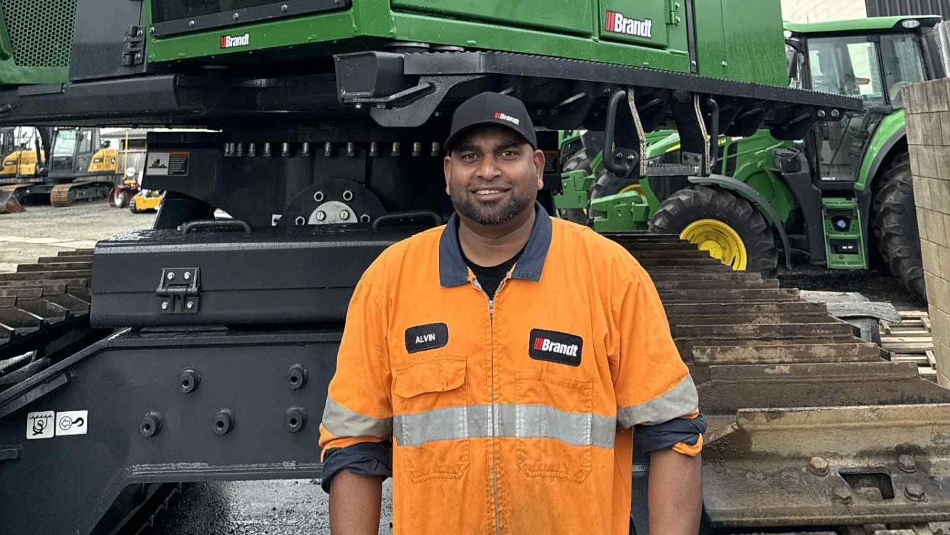A man wearing an orange high visibility shirt and baseball camp in front of a large green forestry machine.