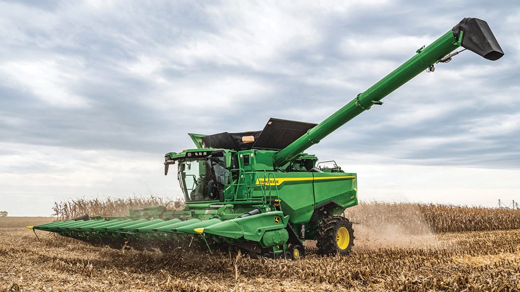 Combine harvester harvesting corn in a field under a cloudy sky, creating dust as it moves along the rows of crops.