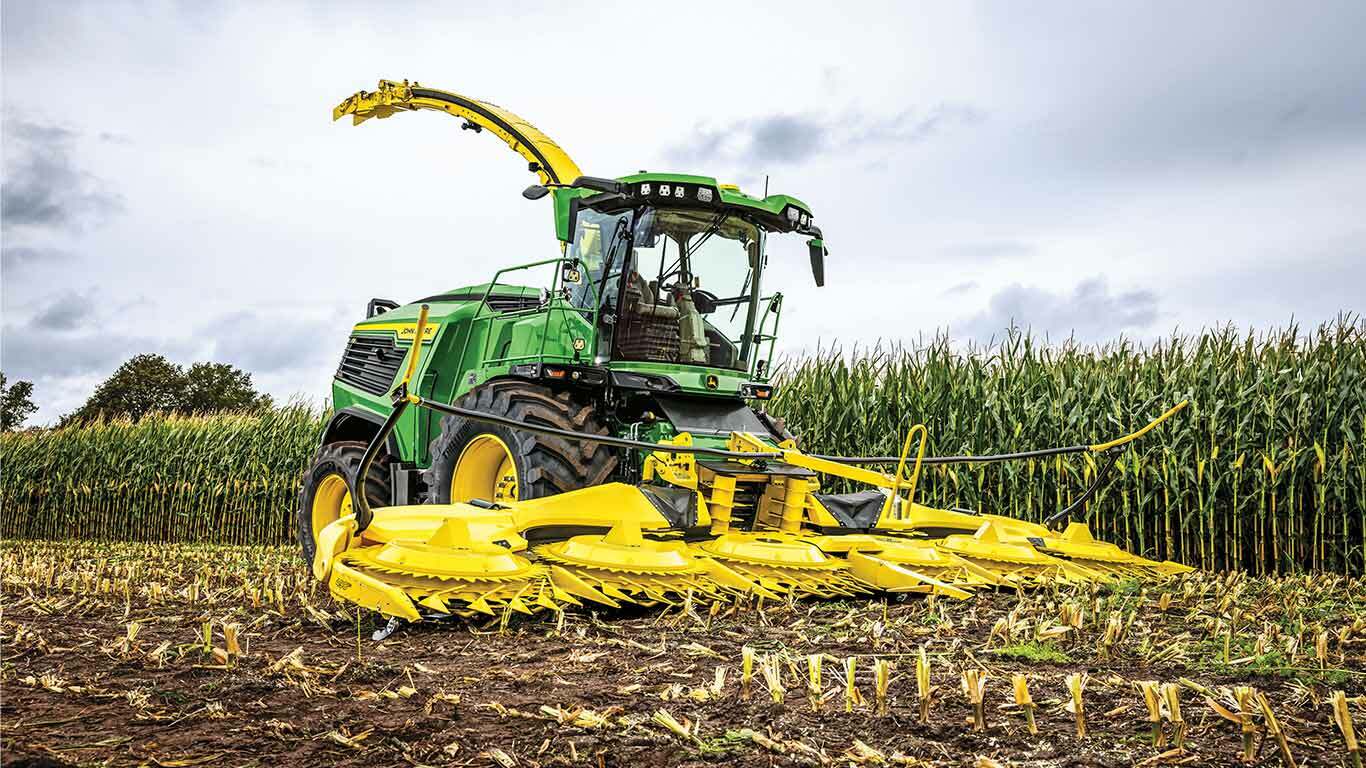 Green and yellow harvester in cornfield with cut stalks in foreground and tall corn behind.