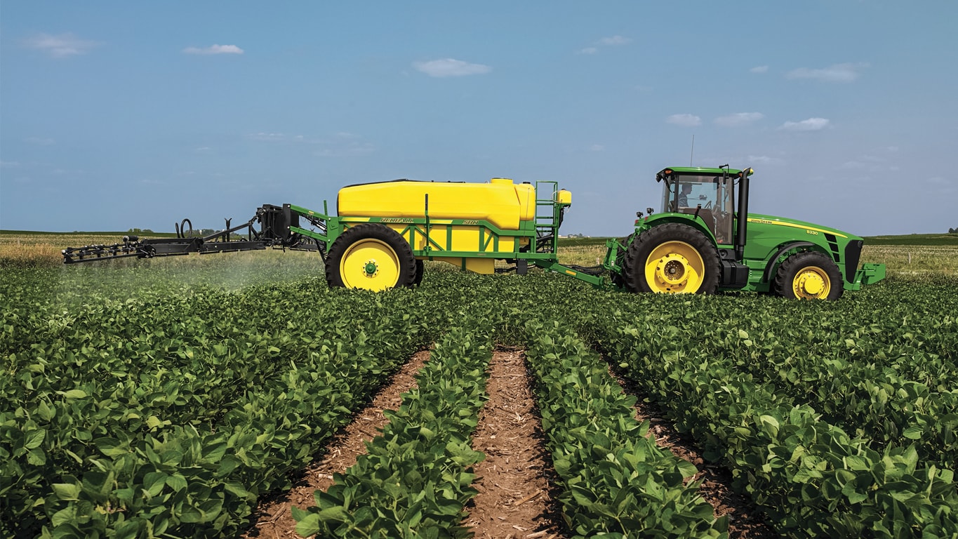 Green tractor with yellow sprayer sprays crops in a large field under a clear blue sky.