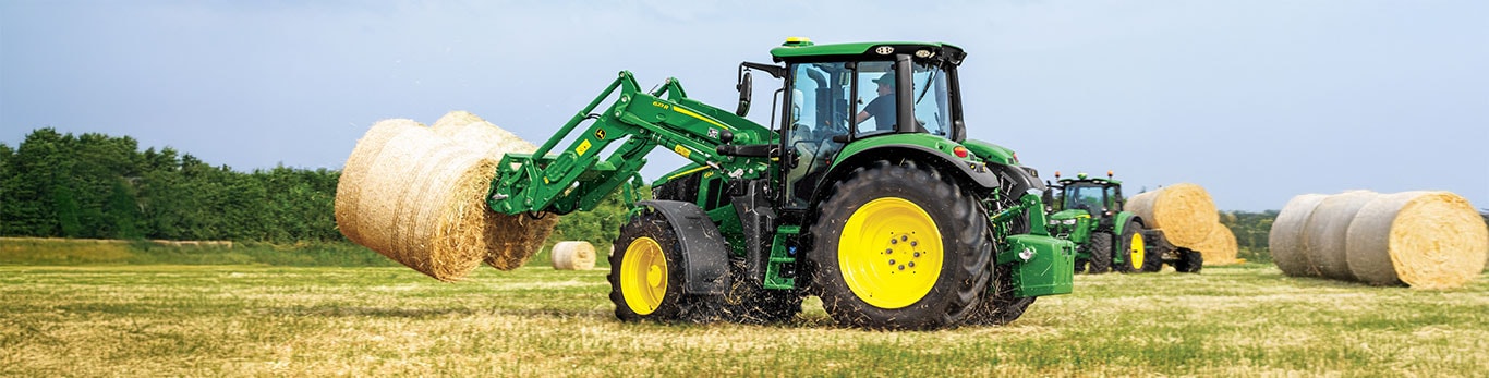 tractor lifts a hay bale in a field with another tractor and hay bales in the background.