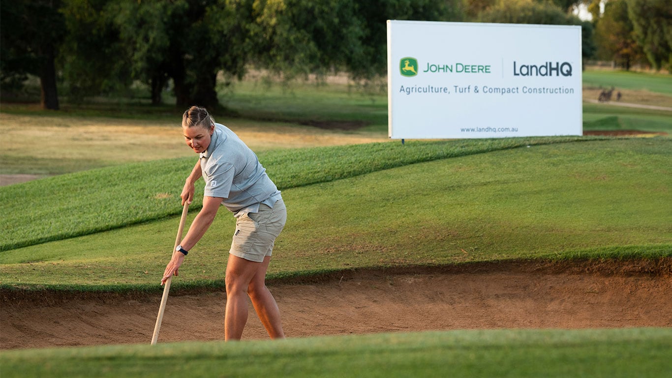 Female Volunteer raking turf in front of John Deere Signage